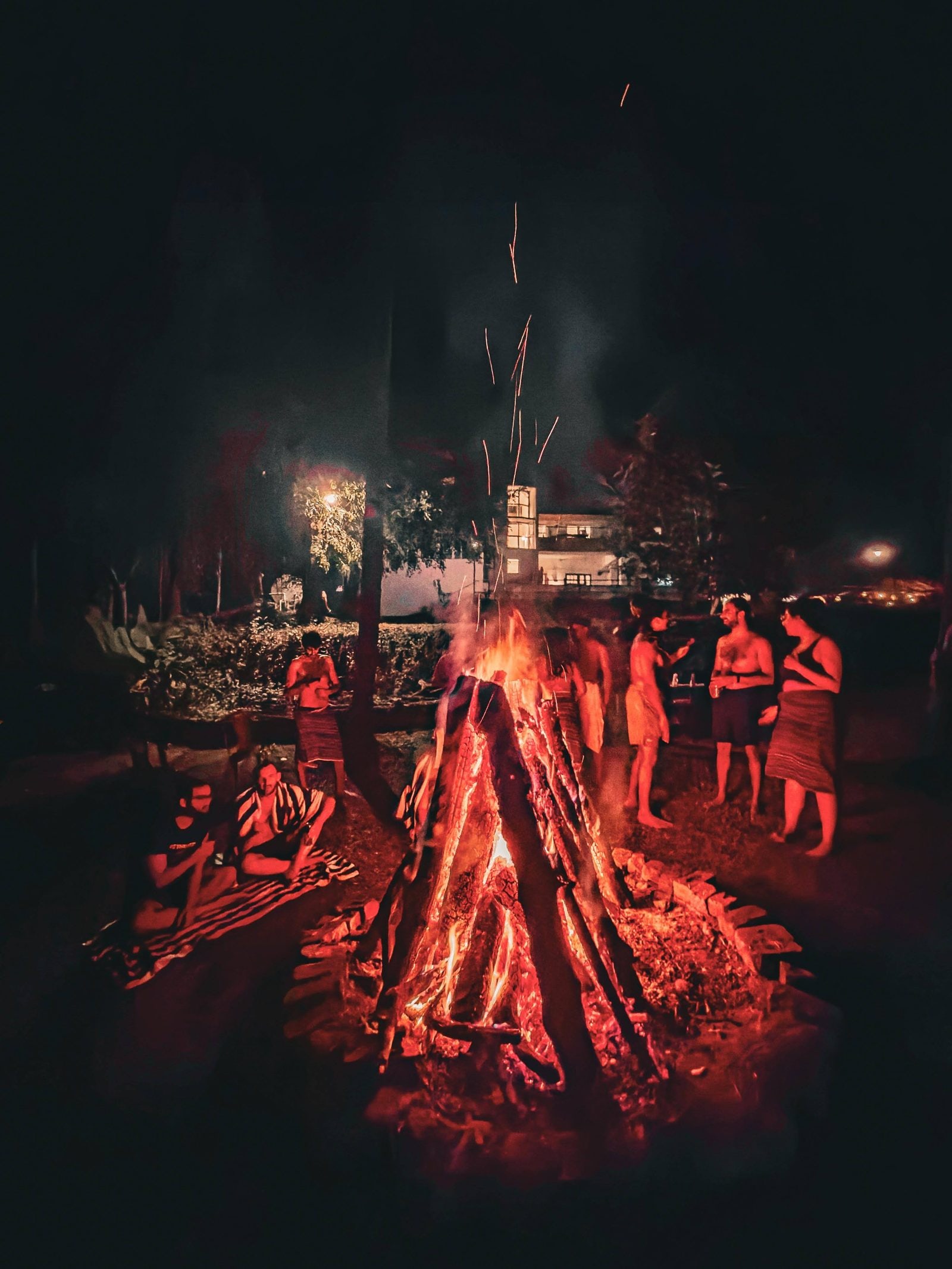 Residents gathered around the bonfire at night, the bungalow glowing behind